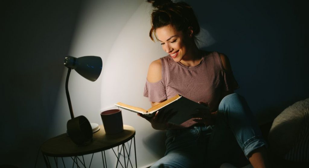 Mulher lendo um livro sob a luz de uma luminária de mesa.